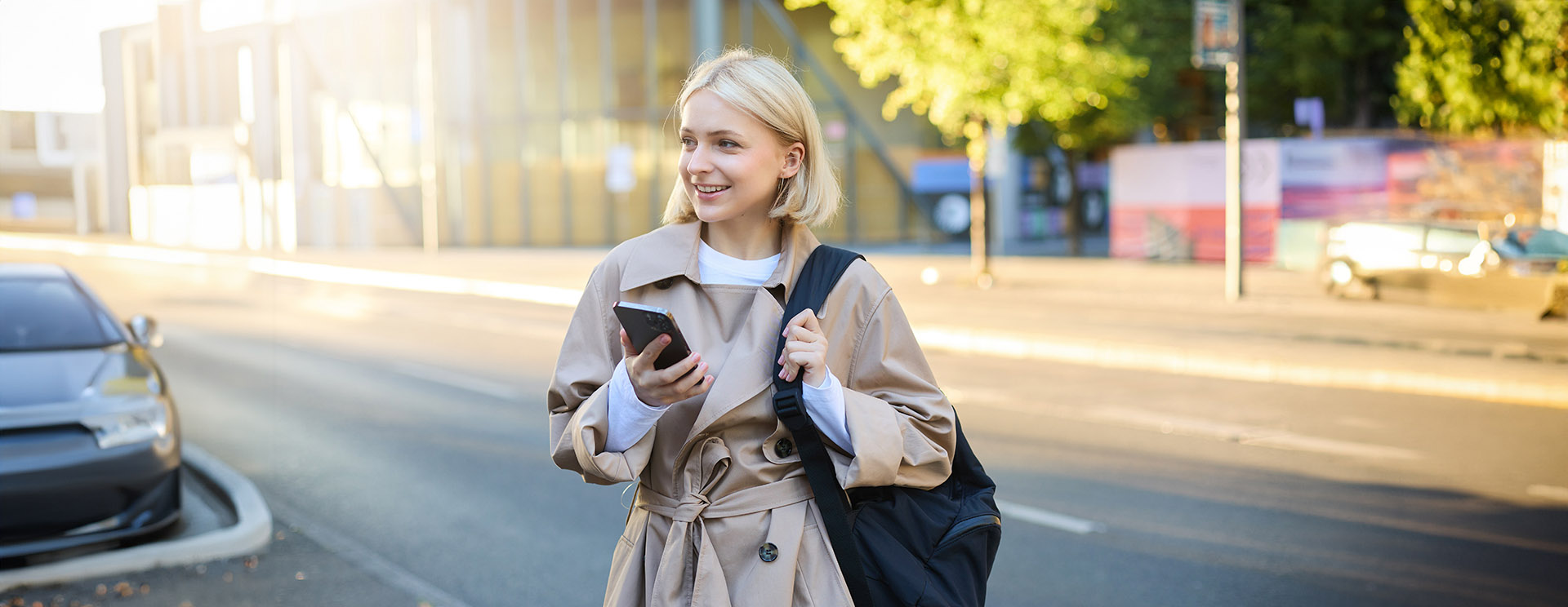 Modern young woman with backpack, student using smartphone, standing on street, sunny day, waiting for ride, using mobile phone application